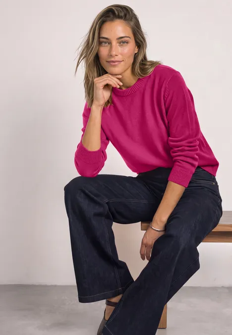 Vrouw in fuchsia trui en wijdere jeans zittend op een bankje, ontspannen voor een lichte achtergrond.