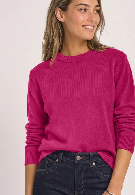 Een lachende vrouw draagt een roze trui en donkerblauwe jeans.
