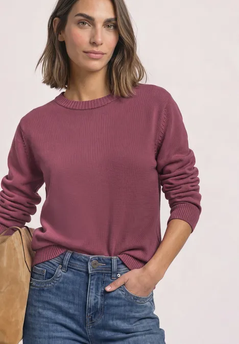 Vrouw in roze trui en blauwe spijkerbroek, ontspannen houding, hand in zak.