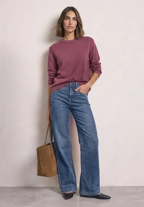 Vrouw voor witte muur, met bordeauxrode trui, wijde jeans en een bruine totebag in de hand.