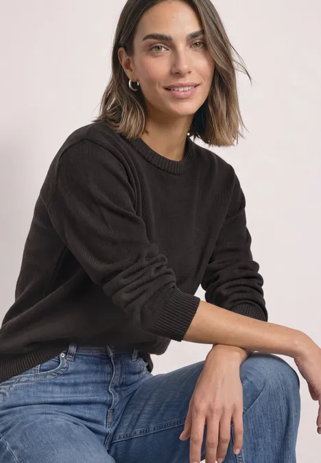 Een lachende vrouw met bruin haar zit ontspannen in een zwarte trui en blauwe jeans tegen een lichte achtergrond.