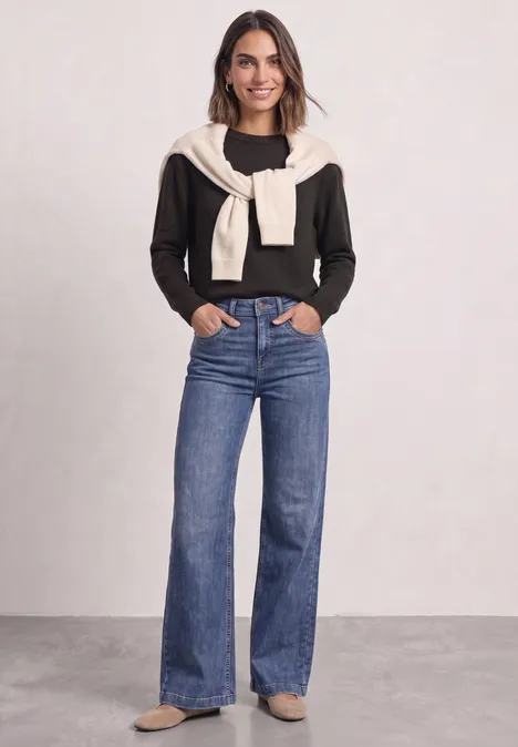Vrouw draagt zwarte trui, beige trui over de schouders en wijde blauwe jeans.