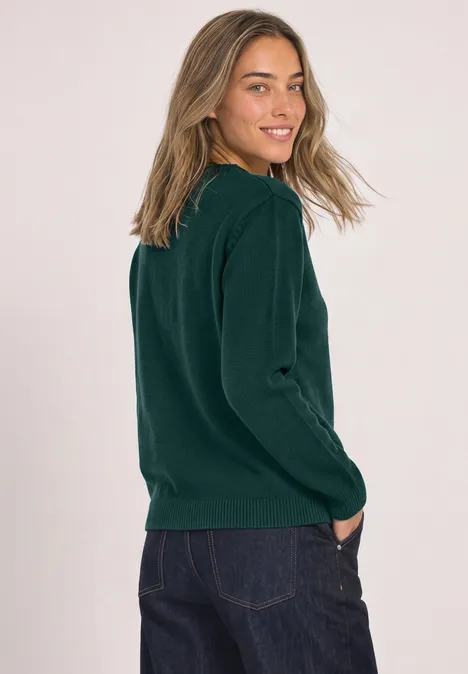 Vrouw met groen trui en spijkerbroek, zijaanzicht, glimlacht naar de camera.
