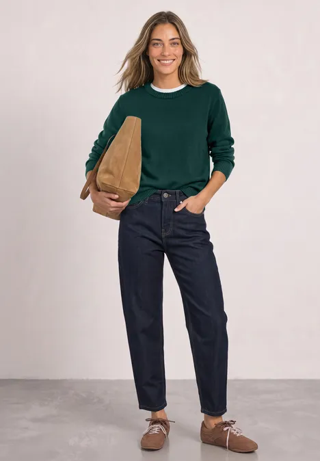 Een vrouw in een groene trui, spijkerbroek en bruine schoenen met een tas glimlachend.