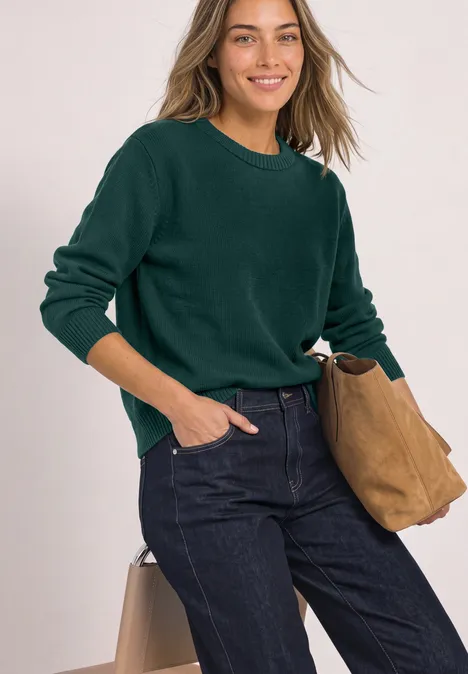 Een jonge vrouw met donkergroene trui, spijkerbroek en beige tas, glimlachend naar de camera.