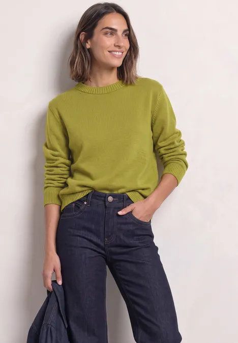 Een vrouw draagt een groene trui en donkerblauwe spijkerbroek, met een jas in haar hand.