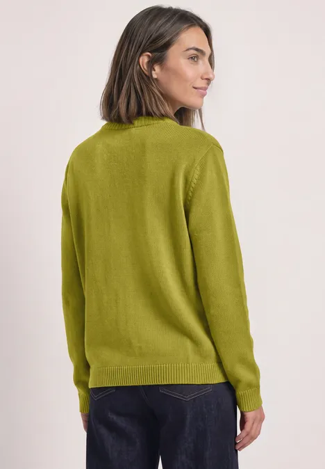 Vrouw in een groene trui, iets gedraaid, kijkend over haar schouder.