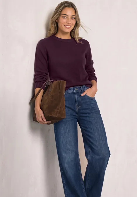 Jonge vrouw glimlachend in paarse trui en wijde jeans, met een bruine tas voor een witte achtergrond.