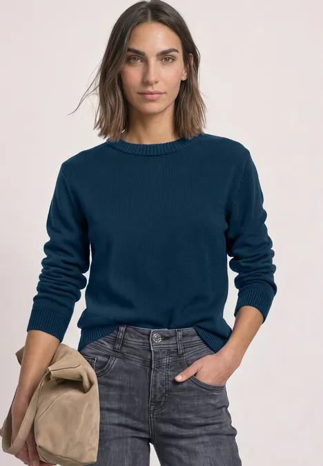 Vrouw in marineblauwe trui en grijze jeans, houdt een beige jas vast, lichte achtergrond.
