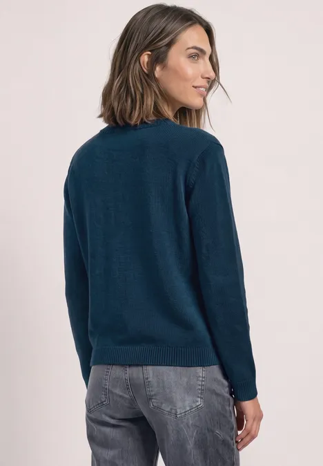 Vrouw in donkerblauwe trui en spijkerbroek, zijwaarts pose, rug licht naar de camera gericht.