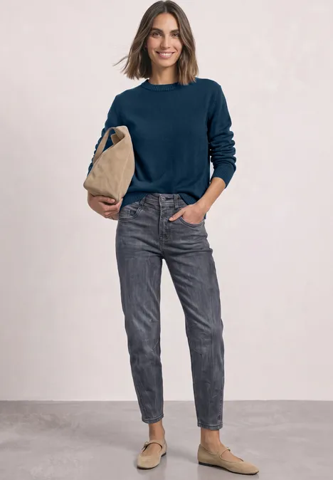 Vrouw met donkerblauw vest, grijze spijkerbroek en beige loafers, met een beige tas voor een neutrale achtergrond.