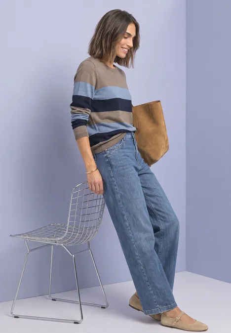 Pullover mit Rundhals und Streifen deep blue Frau in gestreiftem Pullover und weitem Jeans, hält eine Tasche und lehnt an einem Drahtstuhl vor lila Wand.