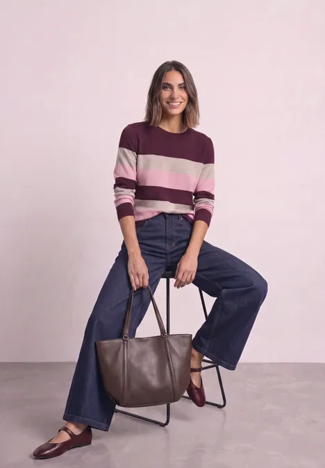 Pullover mit Rundhals und Streifen rustic rouge Frau in gestreiftem Pullover, weiten Jeans und Slippern sitzt lächelnd auf Hocker, hält braune Ledertasche.