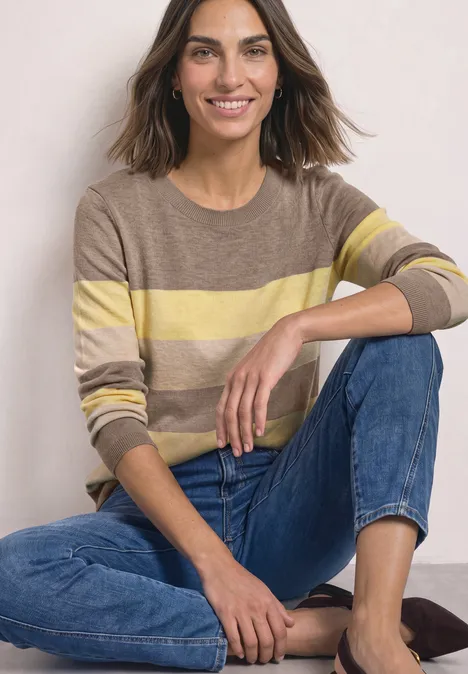 Pullover mit Rundhals und Streifen sanded amber mel. Eine lächelnde Frau in gestreiftem Pullover und Jeans sitzt entspannt auf dem Boden.