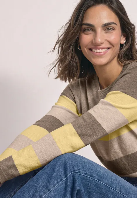 Pullover mit Rundhals und Streifen sanded amber mel. Eine lächelnde Frau trägt einen gestreiften beige-gelben Pullover und Jeans und sitzt entspannt.