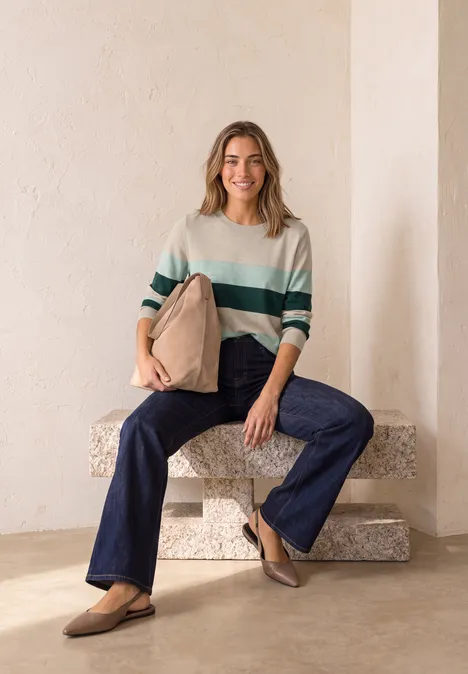 Vrouw zit op een stenen blok, draagt gestreepte trui, wijde jeans en loafers, houdt een beige tas vast.