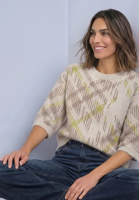 Une femme souriante est assise, portant un pull à manches courtes à motif et un jean, fond pastel.