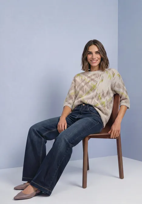 Femme souriante assise sur une chaise, vêtue d’un pull ample et d’un jean, fond neutre.