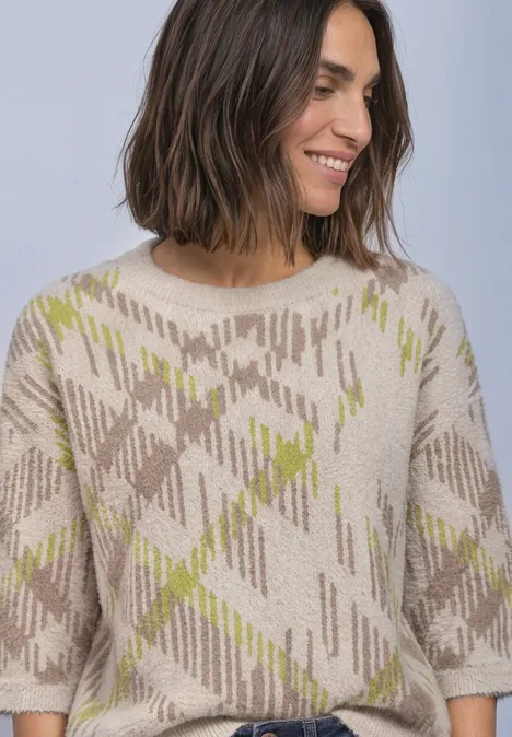 Une femme souriante aux cheveux mi-longs porte un pull beige à motif carreaux verts.