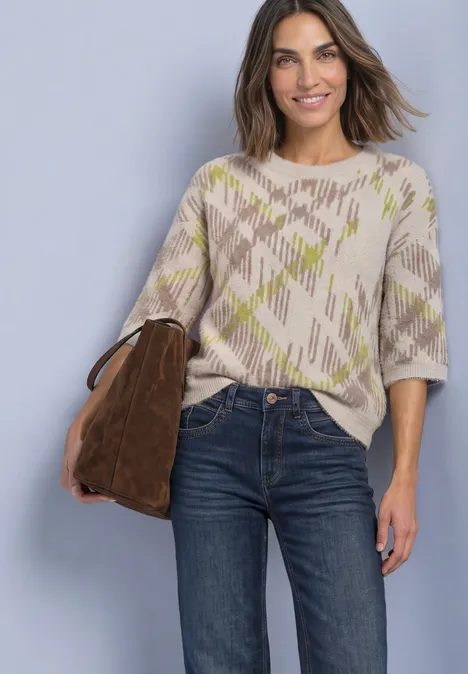 Femme en pull beige à motifs, jean foncé et sac marron souriante devant un mur gris clair.