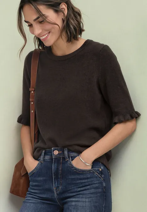 Frau in braunem Shirt mit Rüschenärmeln, Jeans und Tasche lächelt vor hellem Hintergrund.