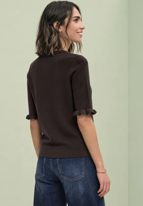 Eine Frau steht mit dem Rücken zur Kamera, trägt einen braunen Pullover mit Rüschenärmeln und blaue Jeans.