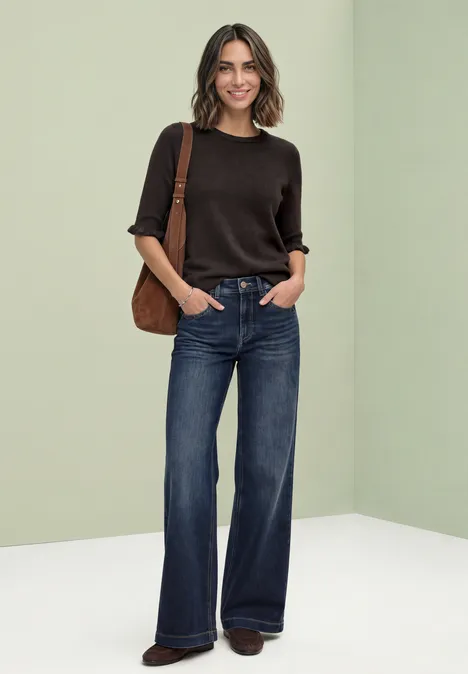 Frau mit braunem Kurzarm-Pullover, weitem Jeans und brauner Schultertasche vor hellem Hintergrund.
