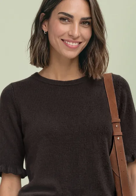 Frau lächelnd in schwarzem Kurzarm-Pullover, trägt eine braune Umhängetasche.