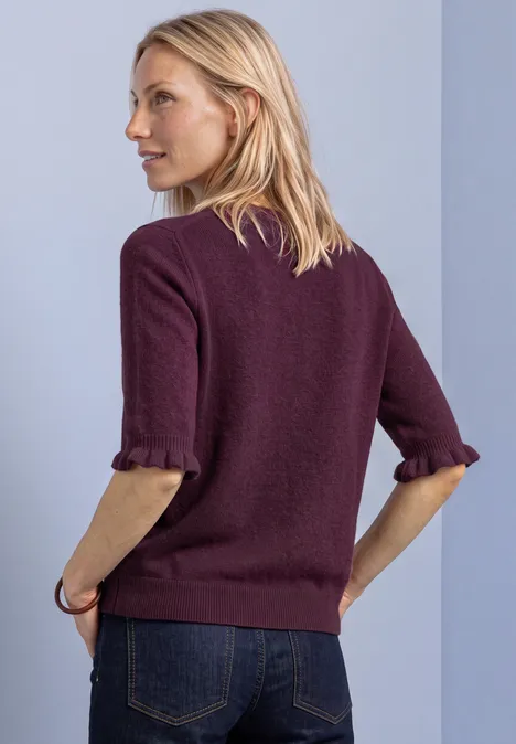 Kobieta z profilu w bordowym swetrze 3/4 z podwiniętymi rękawami oraz dżinsach na jasnym tle.
