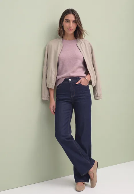 Eine Frau trägt eine rosa Bluse, blaue Jeans und eine Jacke leger über den Schultern.