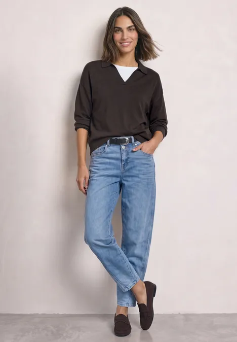 Frau in schwarzem V-Pullover über weißem Shirt, blauen Jeans und schwarzen Loafers vor hellgrauer Wand.