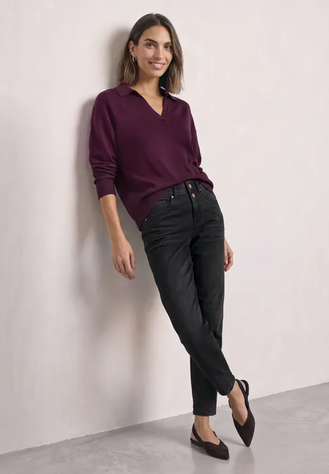 Langarm Pullover mit Polokragen berry maroon Eine lächelnde Frau in violettem Pullover lehnt locker an einer Wand, trägt schwarze Jeans und flache Schuhe.
