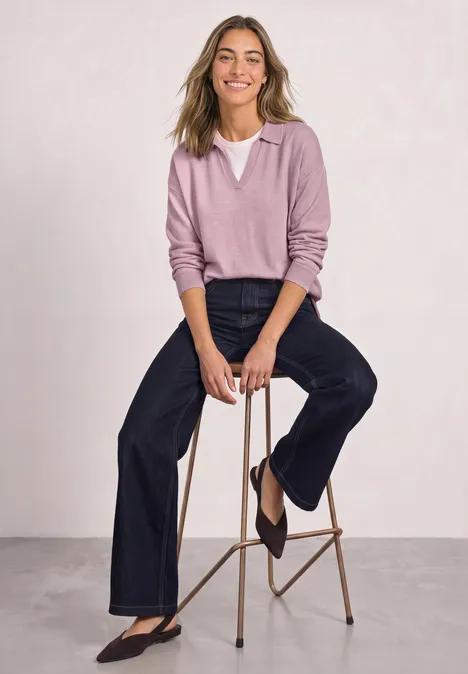 Frau sitzt auf Hocker, trägt rosa Pullover mit V-Ausschnitt und weite Jeans, lächelt entspannt.