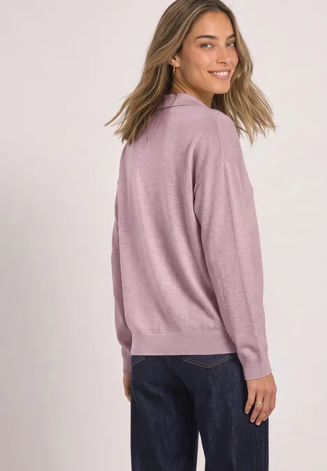 Frau in rosa Pullover und dunklen Jeans blickt lächelnd über die Schulter vor weißem Hintergrund.