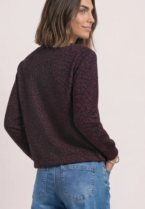 Seitlich zugewandte Frau mit schulterlangen Haaren trägt einen lila Pullover mit Leopardenmuster und blaue Jeans.