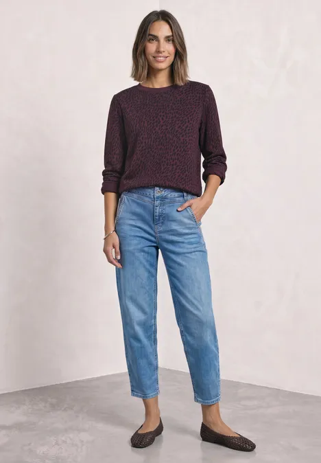 Eine Frau trägt burgunderfarbenen Pullover, blaue Jeans und braune Loafers vor hellem Hintergrund.