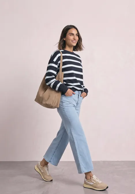 Frau in gestreiftem Pullover und Jeans mit Tragetasche, die lächelnd einen Raum betritt.