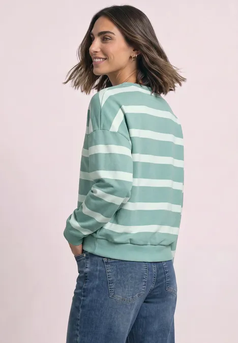 Frau in grün-weiß gestreiftem Sweatshirt und blauer Jeans, die seitlich steht und lächelnd zurückblickt.