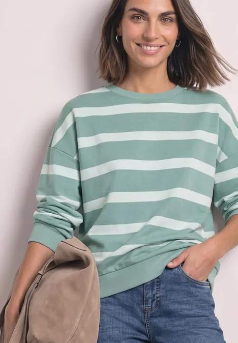 Frau in grün-weiß gestreiftem Pullover, Jeans, lächelt, trägt eine Tasche.