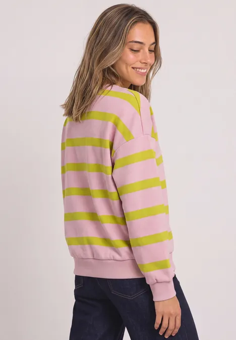 Frau im rosa-grün gestreiften Pullover und dunkler Jeans, seitlich stehend und lächelnd.