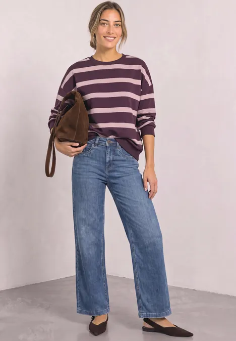 Frau in lila-gestreiftem Pullover, weiten blauen Jeans und schwarzen Schuhen, mit brauner Tasche vor heller Wand.