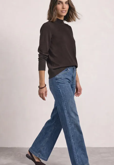 Vrouw in wijde blauwe jeans en een bruine trui staand zijwaarts voor een lichte achtergrond.
