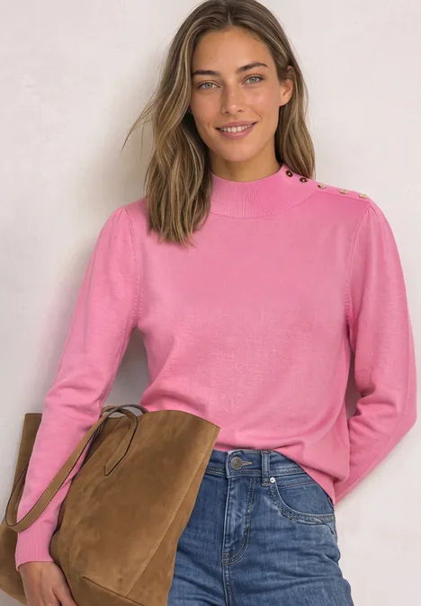 Eine lächelnde Frau trägt einen rosa Pullover und Jeans, eine braune Tasche über der Schulter.