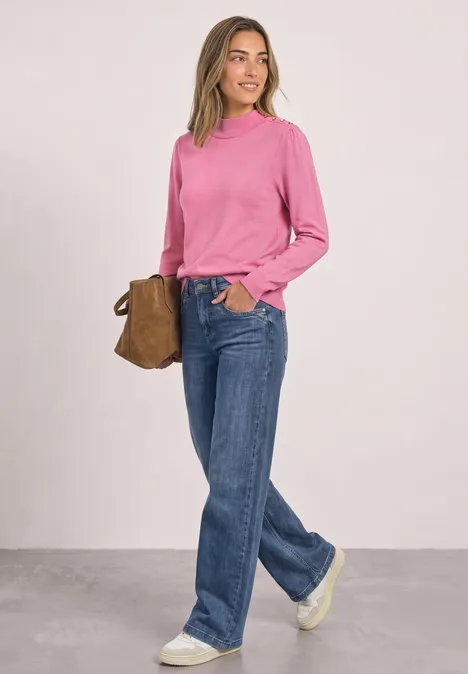 Junge Frau trägt pinken Pullover, blaue Jeans und weiße Sneaker, hält eine braune Tasche.