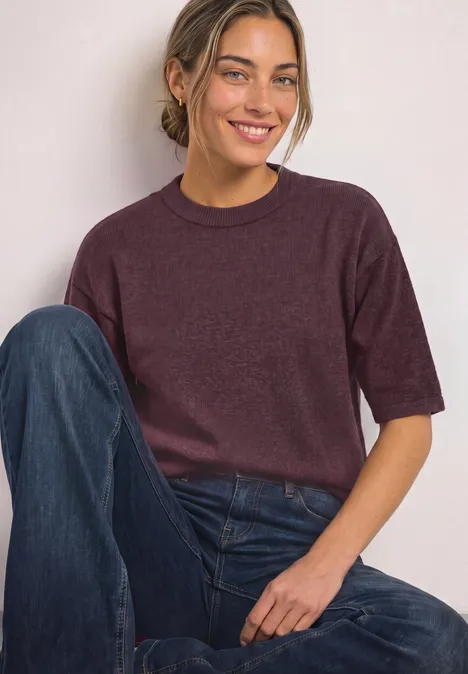 Junge Frau sitzt lässig, trägt lila Pullover und blaue Jeans, lächelt entspannt in die Kamera.