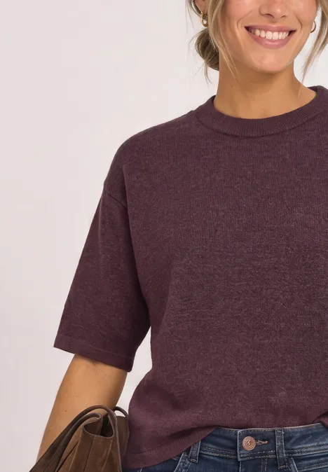 Frau mit weinrotem T-Shirt und Jeans, lächelt, hält braune Tasche.