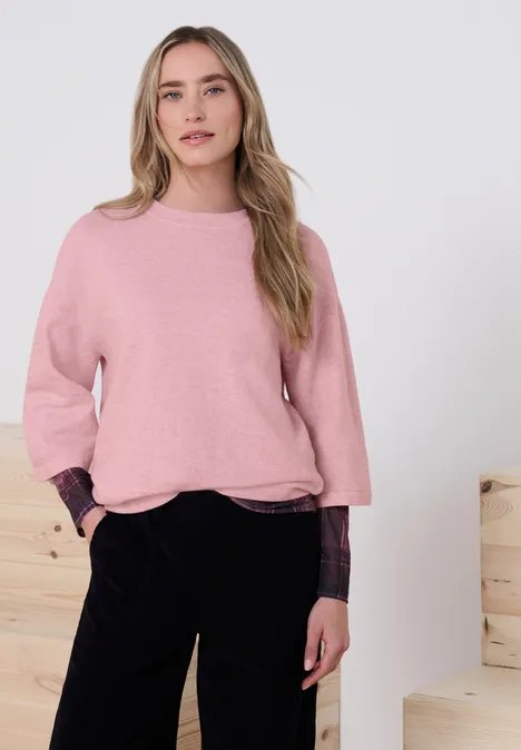Jonge vrouw draagt een roze trui en zwarte broek, handen in de zakken, voor lichte houten blokken.