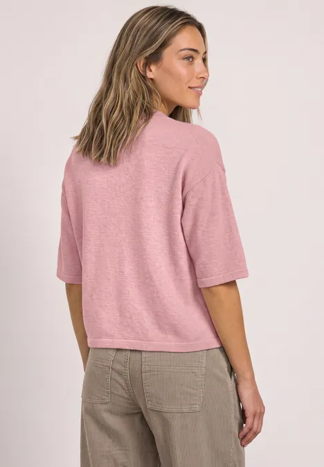Een vrouw in een roze T-shirt en beige broek staat licht van opzij en kijkt opzij.