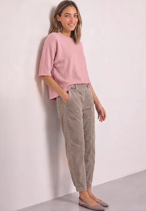 Een glimlachende vrouw leunt tegen een muur, draagt een roze top, grijze ribfluwelen broek en grijze ballerina’s.