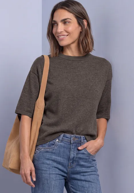 Eine Frau lächelt, trägt ein braunes Kurzarmshirt, blaue Jeans und eine beigen Stofftasche.
